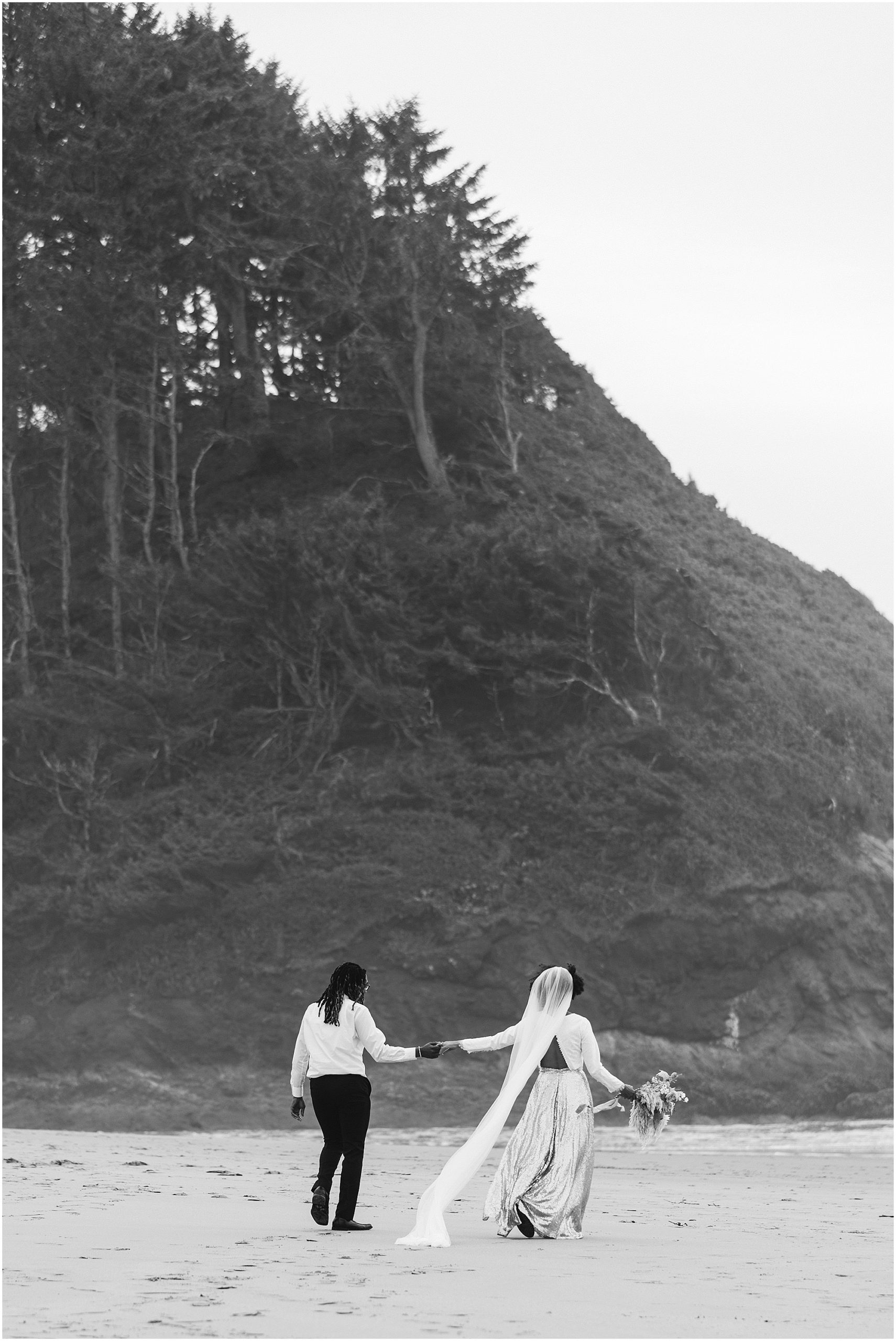 A gorgeous black and white image of a couple walking in the sand in their wedding attire holding hands after their intimate Oregon Coast elopement near Lincoln City. | Erica Swantek Photography