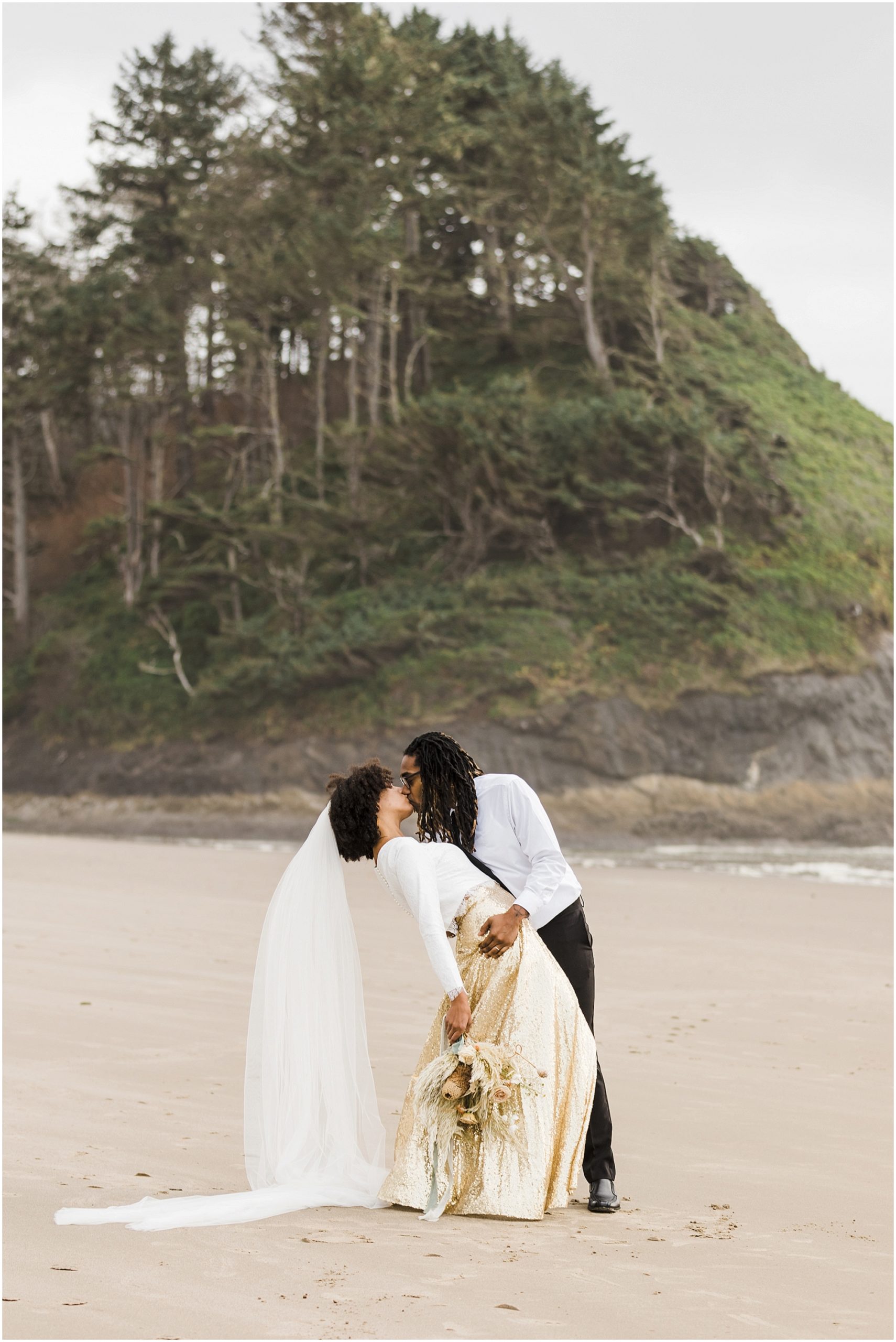 A groom wearing a white shirt and black pants dips his bride wearing a gold sequined skirt back for a kiss with Proposal Rock in the background. They married in an intimate ceremony on the Oregon Coast. | Erica Swantek Photography
