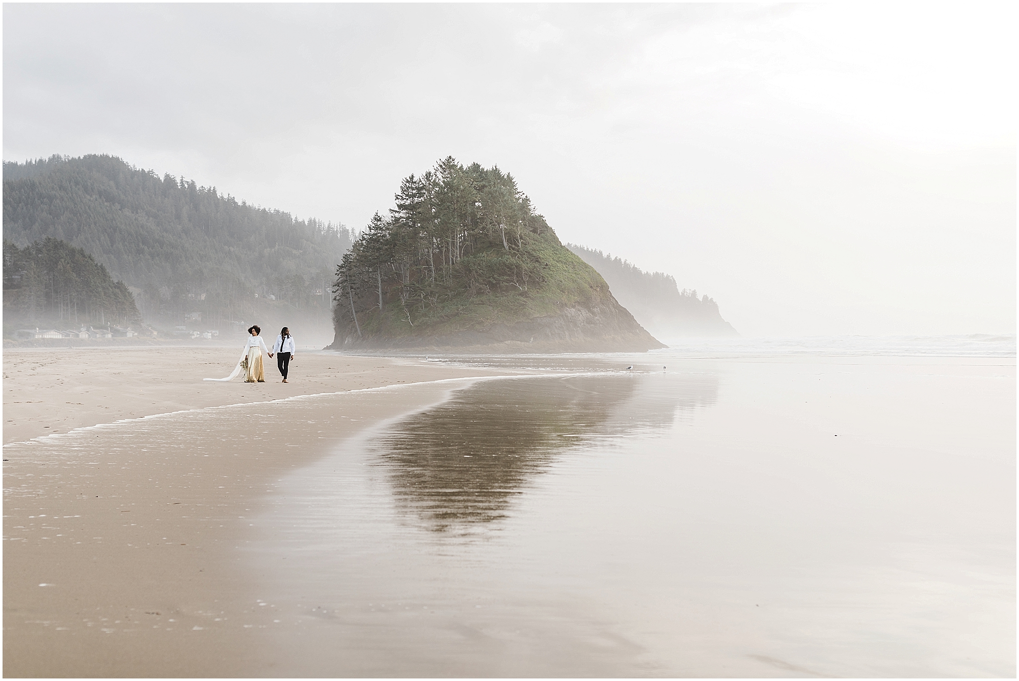The Pacific Ocean in the foreground reflects Proposal Rock as a gorgeous black bride and room walk hand in hand along the sand during their Oregon Coast elopement. | Erica Swantek Photography 