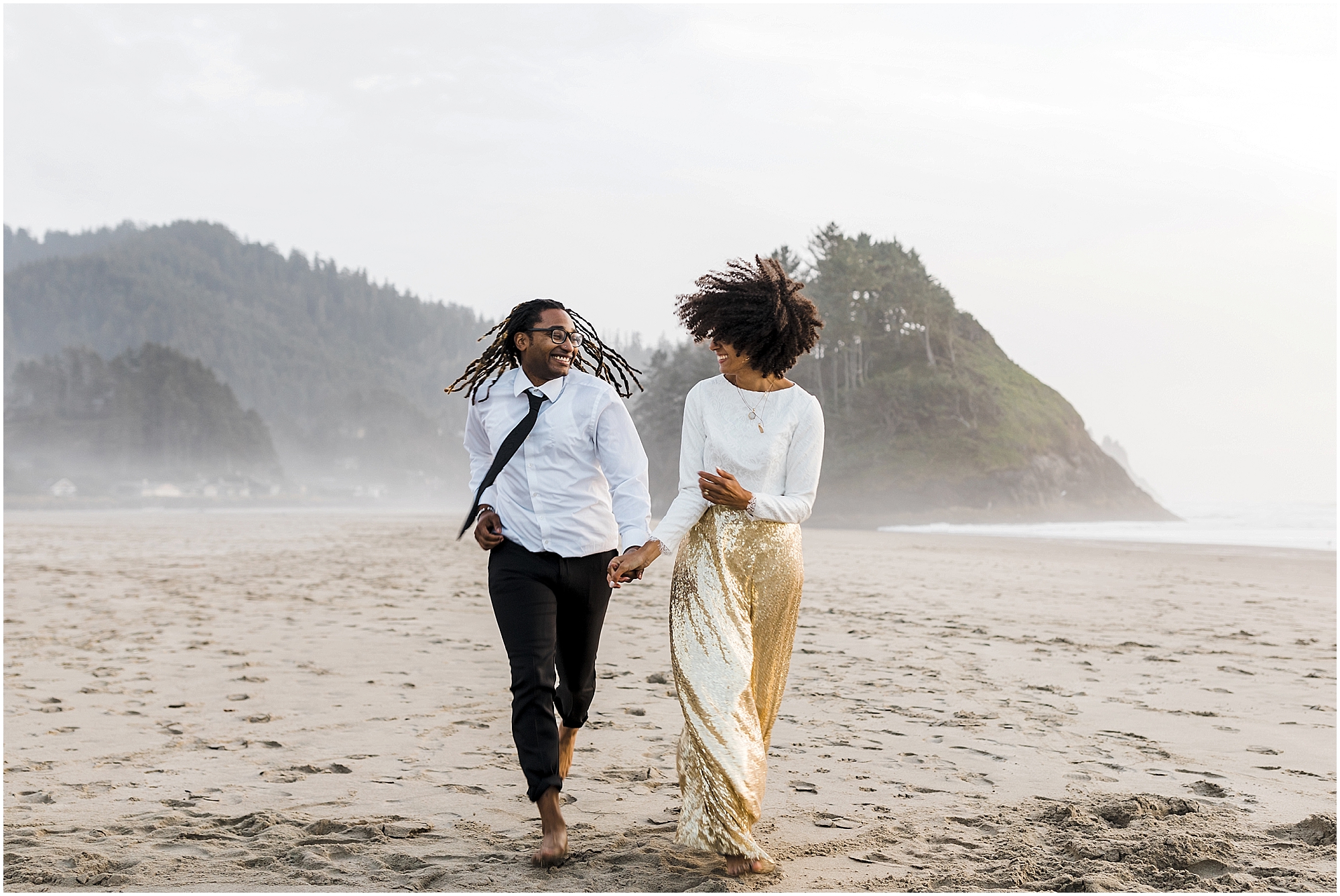 A black couple runs hand in hand in the sand in their wedding attire with the rugged Oregon coastline behind them for their beach elopement near Lincoln City. | Erica Swantek Photography