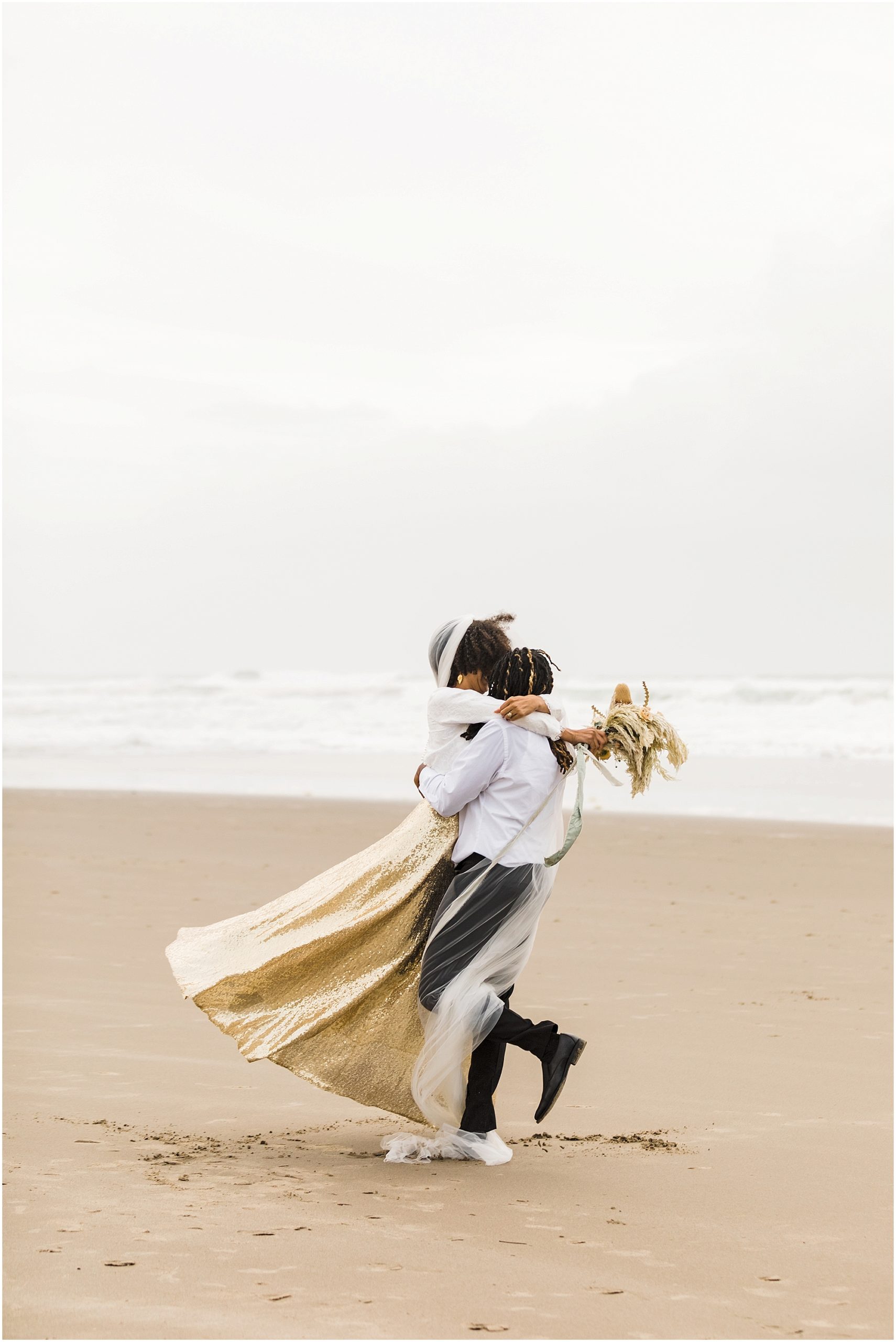 A groom spins his gorgeous black bride in his arms as her gold sequined skirt and long white veil flies in the wind. She tosses her bouquet over his shoulder with the Pacific Ocean behind her. | Erica Swantek Photography