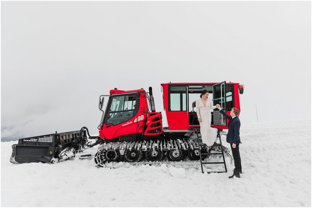 A groom wearing a blue tuxedo jacket and black bowtie takes the hand of his bride wearing a long sleeve laced gown as he helps her down the black iron ladder from the red snowcat machine for their Silcox Hut intimate winter wedding at Timberline Ski Resort in Oregon.