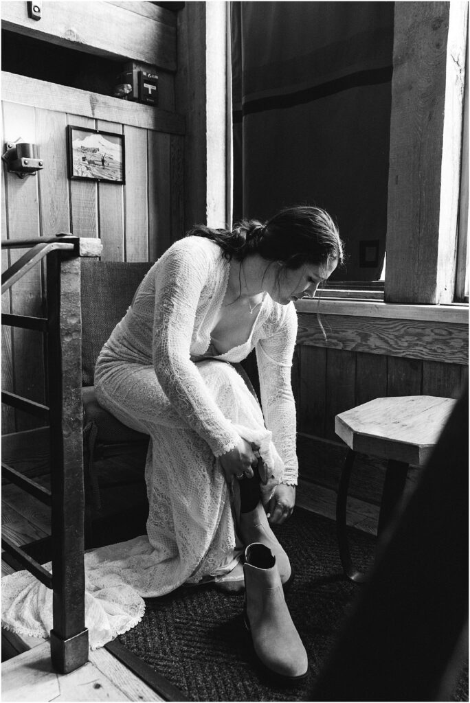 A black and white image of a bride sitting next to a window in her long sleeved lace wedding gown as she puts on her Sorel boots before seeing her groom outside of the Silcox Hut for their wedding day. 