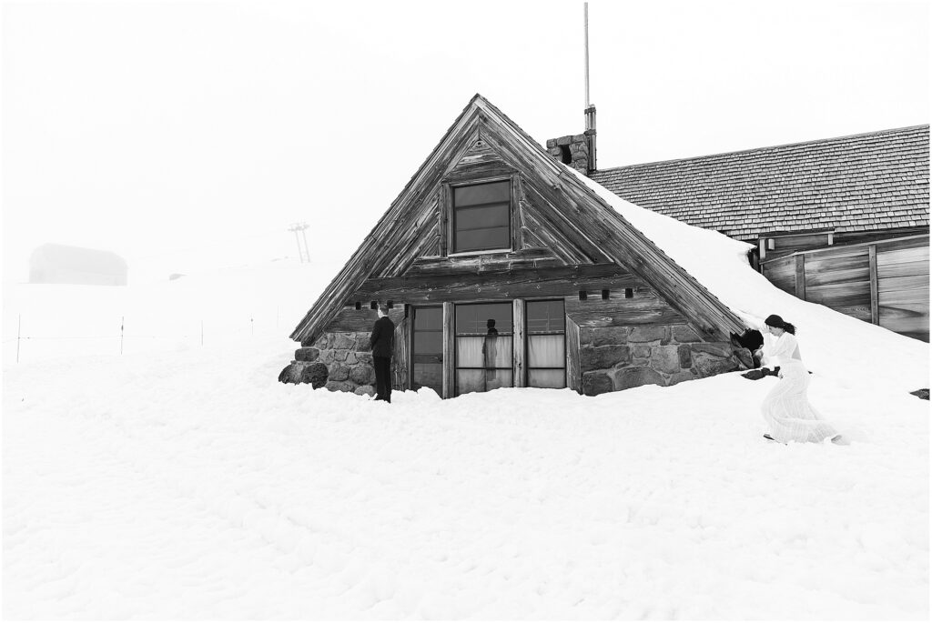 A black and white image of a bride wearing a white lace gown walking through the snow towards her groom with his back turned. They are standing in front of the Silcox Hut on Mt. Hood. 