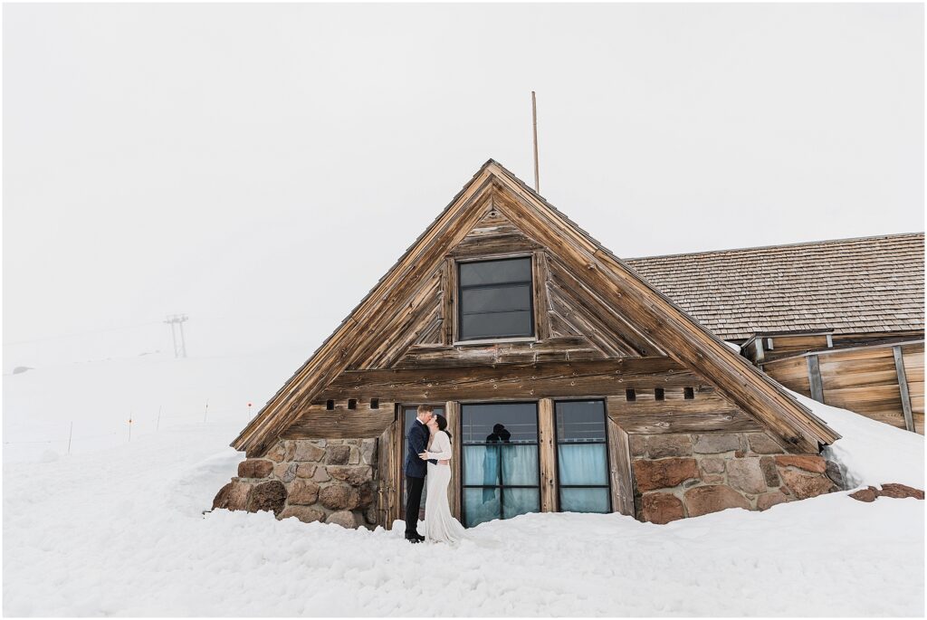 A bride and groom embrace and kiss in front of the Silcox Hut for their winter wedding in the snow. 