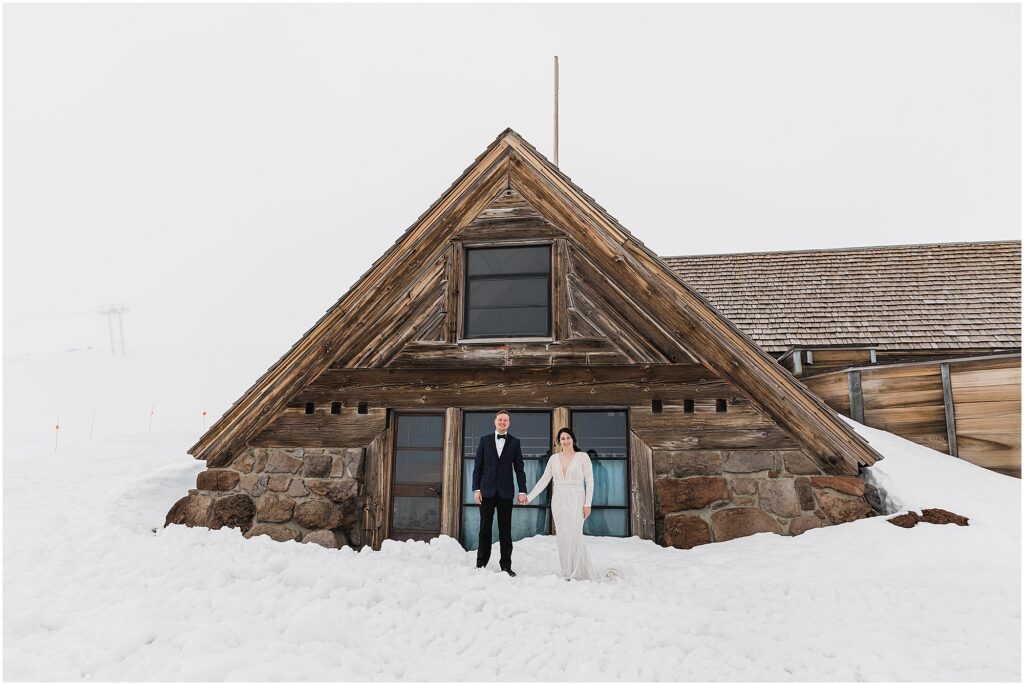 A bride in a white lace gown with long sleeves holds her groom's hand wearing a blue jacket and black bowtie in front of the rustic wood and rock Silcox Hut Lodge at Timberline Ski Resort for their Oregon winter ski elopement.