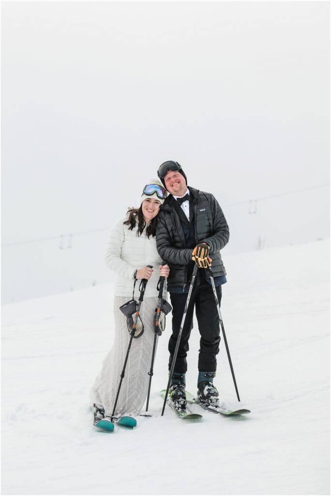 A bride and groom stand in their wedding clothes wearing skis on Mt Hood for their Silcox Hut intimate winter wedding. 