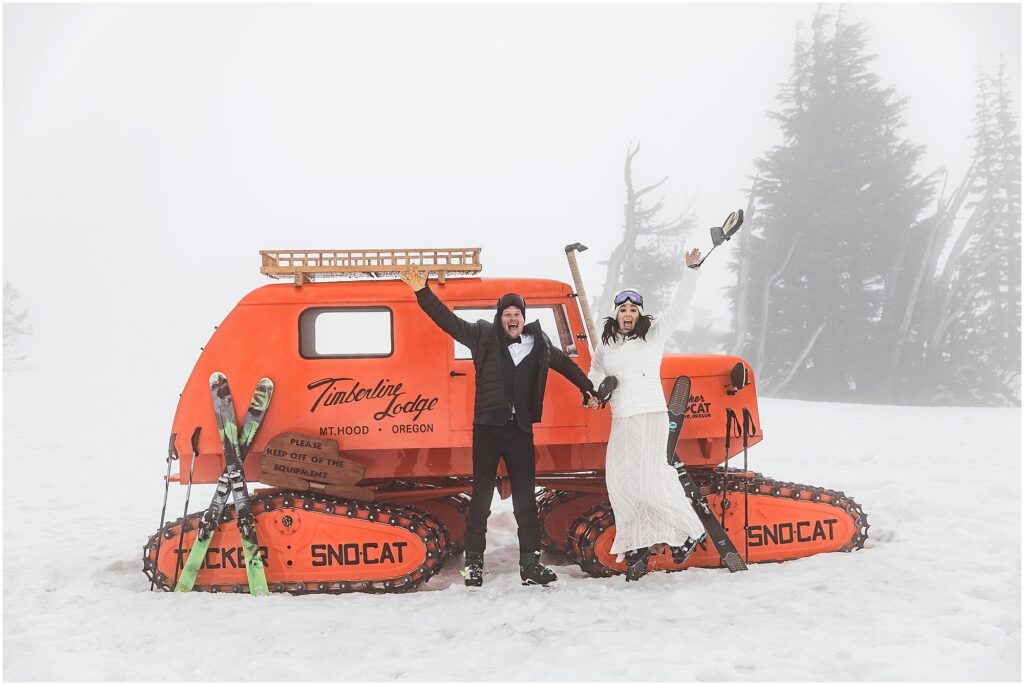 A bride is a white dress, ski boots, white jacket and googles umps in the air while holding her groom's hand. He is in a black suit with black jacket wearing ski boots and googles. They stand in front of a vintage orange snowcat machine with Timberline Lodge, Mt. Hood, Oregon written the side.