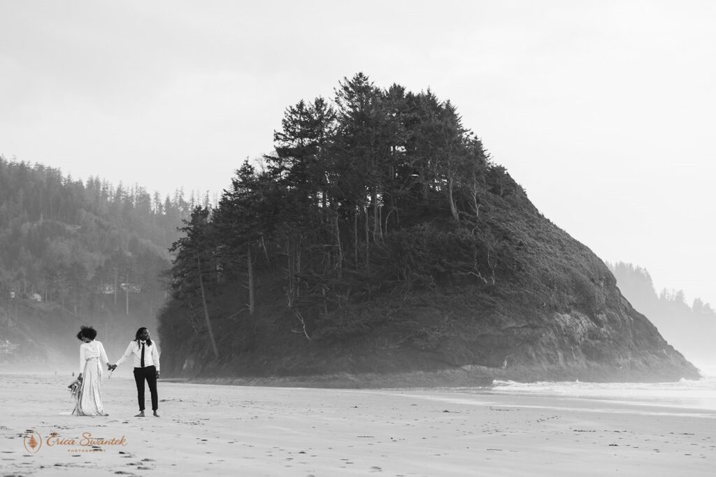 A couple is seen in the distance walking near a large rock formation on an Oregon beach.
