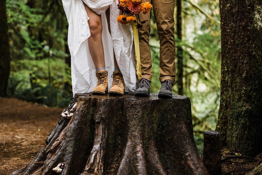 A couple stands on a tree stumps and shows their hiking boots.