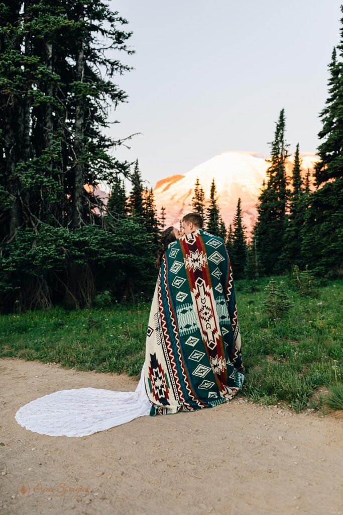 A couple cuddles underneath a blanket while kissing in front of Mt. Rainier.