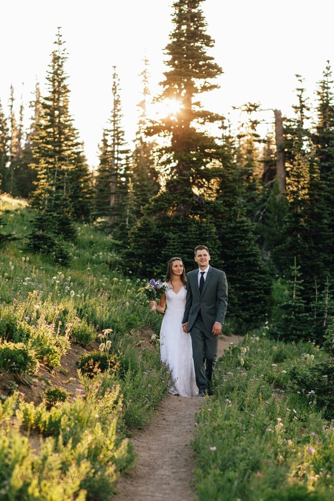 An adventure elopement couple walks along a trail near a meadow of wildflowers.