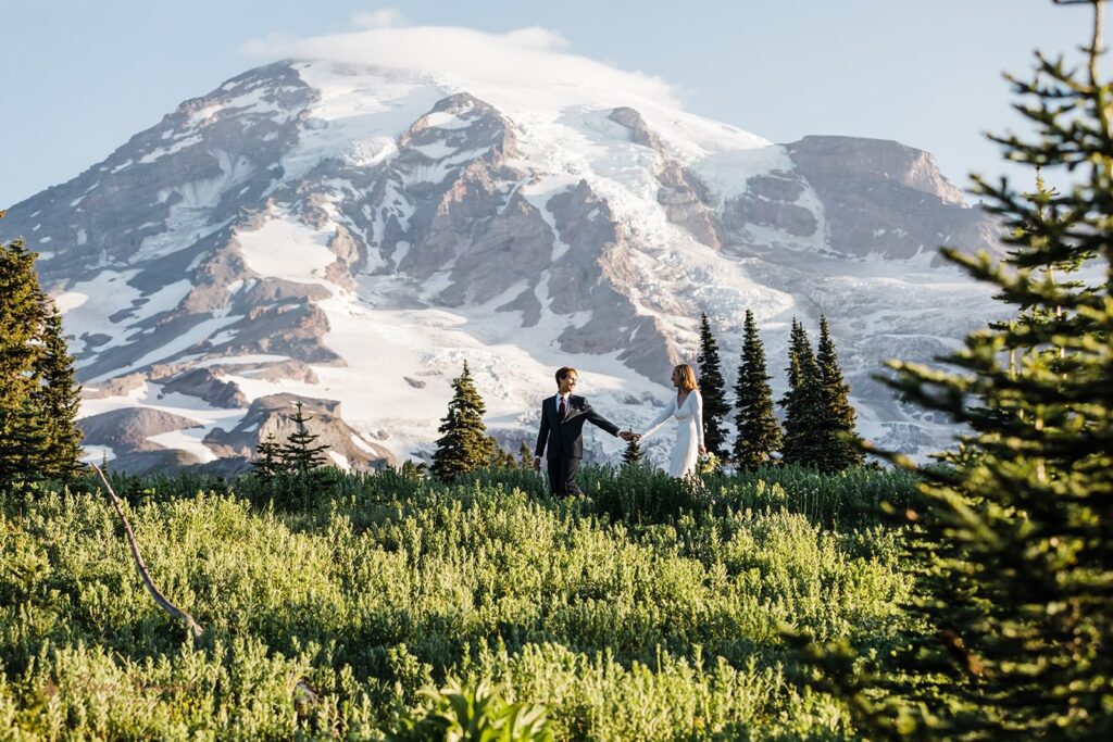 A couple holds hands while walking through a meadow beneath Mt. Rainier. 