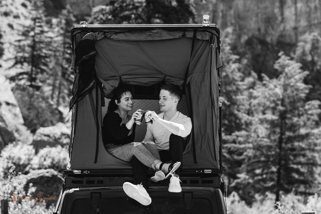 An engaged couple cheers drinks while sitting in a rooftop tent. 