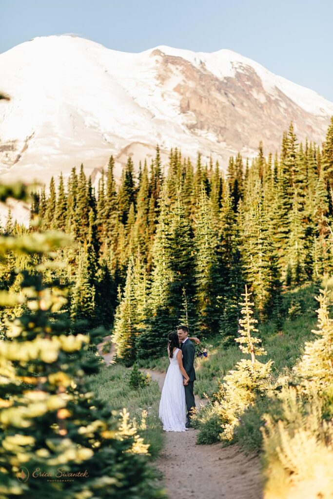 A couple stands along a dirt trail beneath the base of Mt. Rainier while wearing wedding attire. 