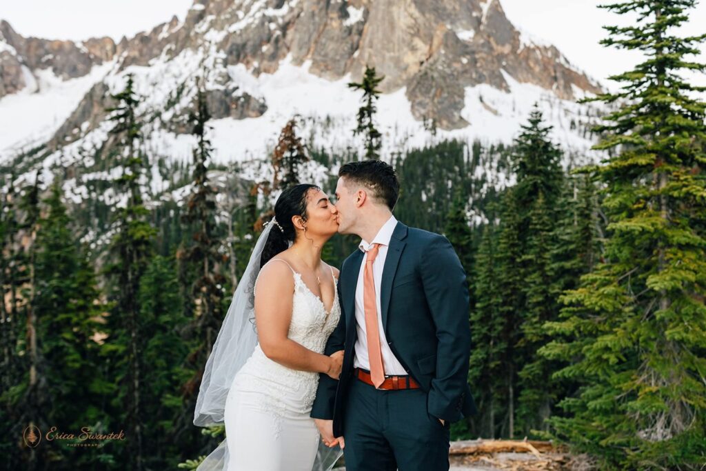 A newlywed couple kisses in an evergreen forest.
