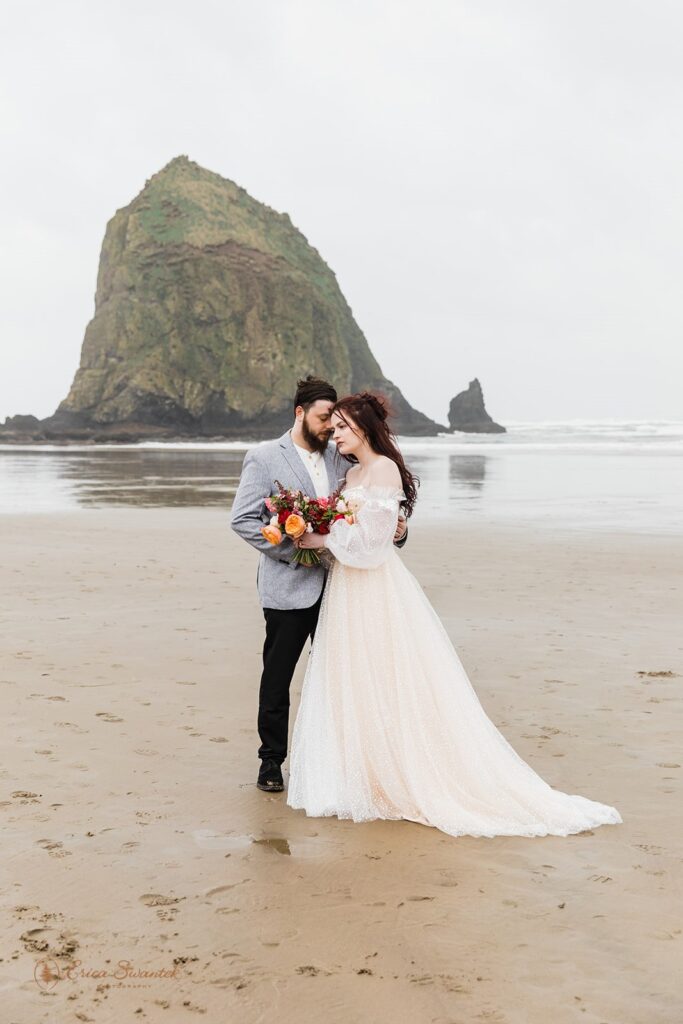 A bride is seen wearing a boho wedding dress on Cannon Beach with her groom who is wearing a grey suit coat. 