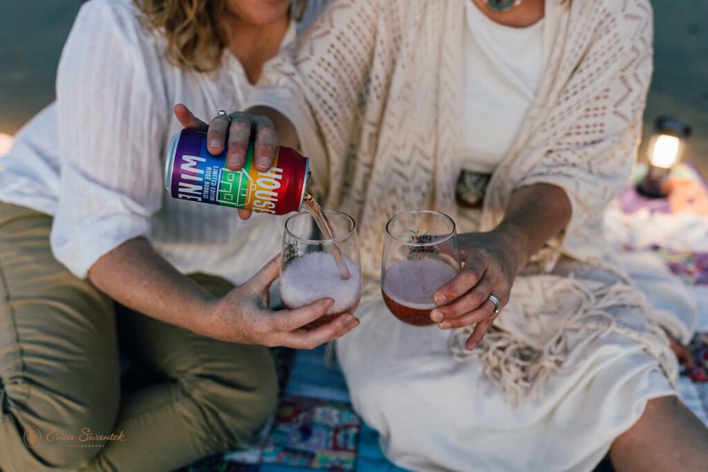 A couple shares a glass of rainbow wine each on the beach during a Sunset elopement picnic. 
