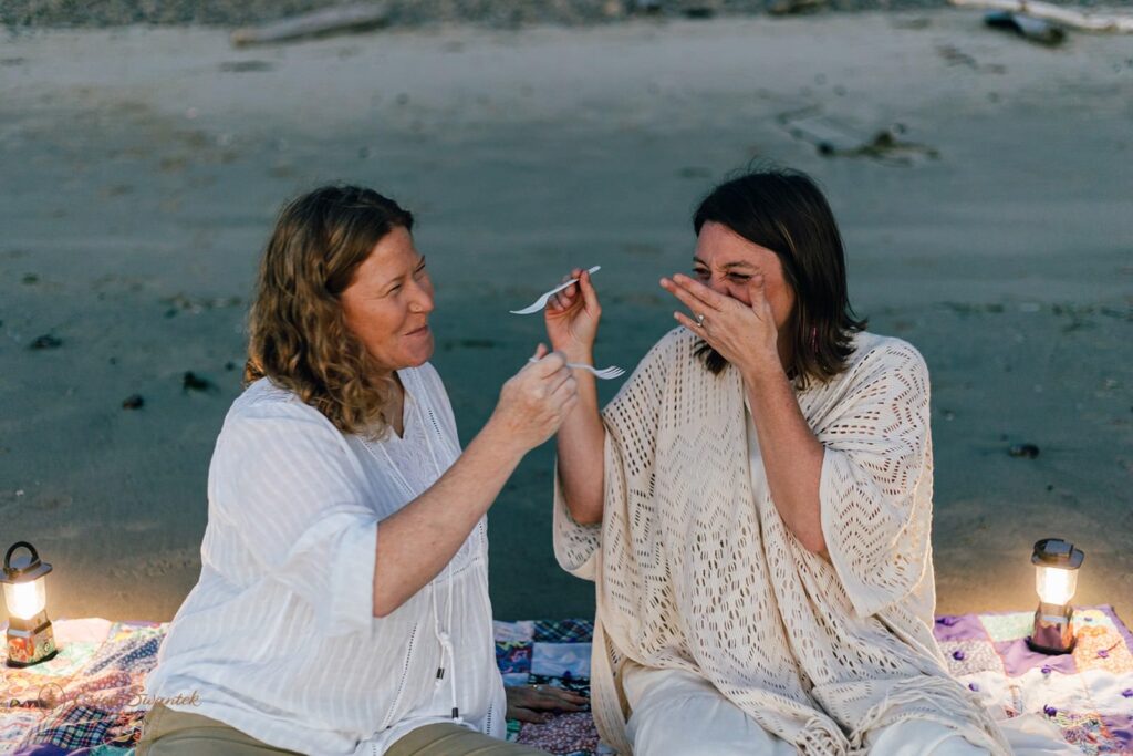 A couple shares bites of wedding cake at the beach. 