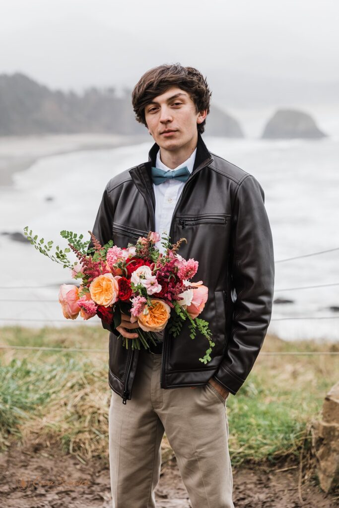A man, on an Oregon Coast overlook, holding a colorful bridal bouquet is wearing a leather jacket, khaki pants, blue bowtie and white dress shirt. 