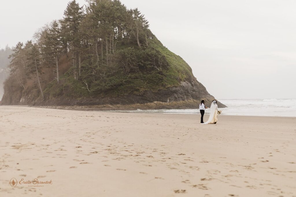 A groom and bride walk along a sandy beach in Oregon near Proposal Rock.