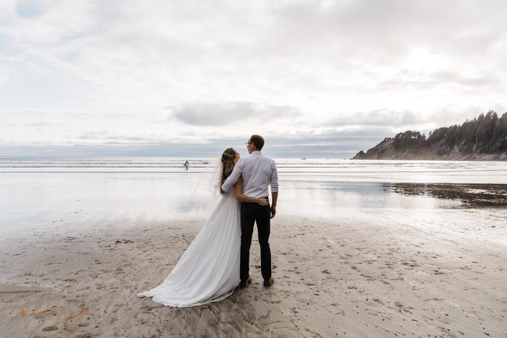 A bride in a long, white wedding gown and veil hugs her groom as they lookout onto the Pacific Ocean during their Oregon Coast elopement.