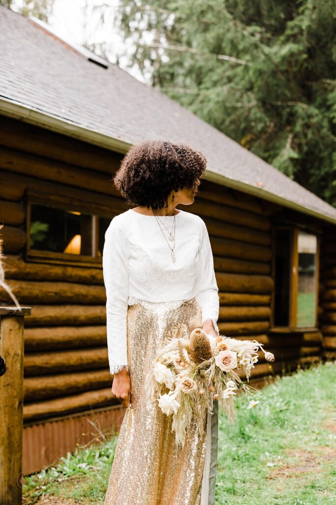 A bride wears a sparkly skirt and white, long sleeve blouse, while holding neutral wedding florals outside an Oregon Airbnb.