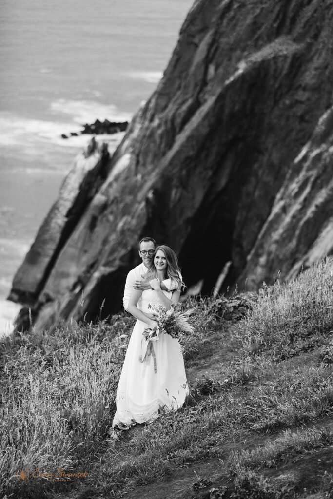 A bride holds a wildflower bouquet while her groom holds her in a coastal meadow overlooking the Pacific Ocean.