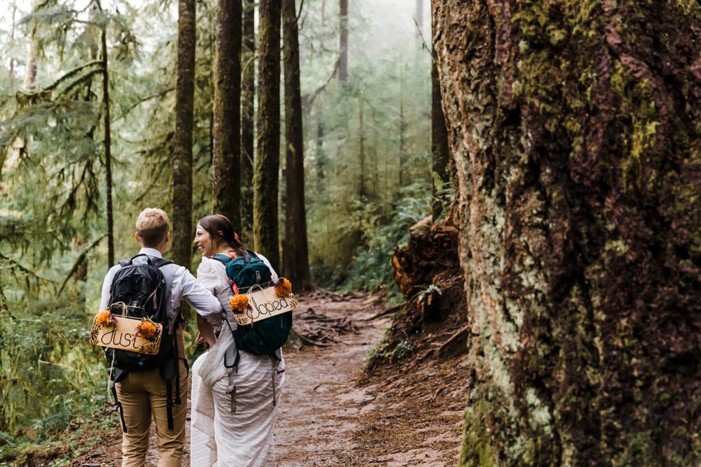 A couple walks along a hiking path in an Oregon forest wearing just eloped signs on backpacks. 