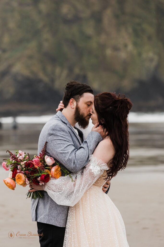 A woman holds wedding florals while kissing her husband during elopement portraits at Cannon Beach.