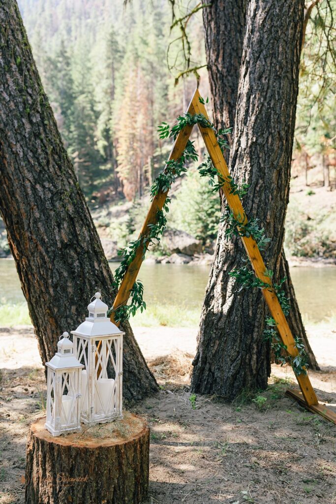 Two white wedding lanterns on a tree stump near an a-frame arch decorated with greenery. 