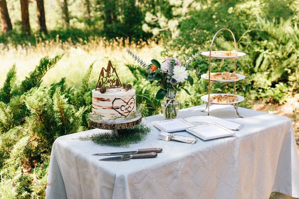 A rustic cake table with a one-tier wedding cake, colorful florals in a vase, and a three-tiered dessert tray. 