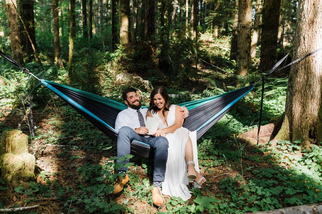 A couple laughs together while sitting in a hammock in an evergreen forest near Mt. Rainier National Park. 