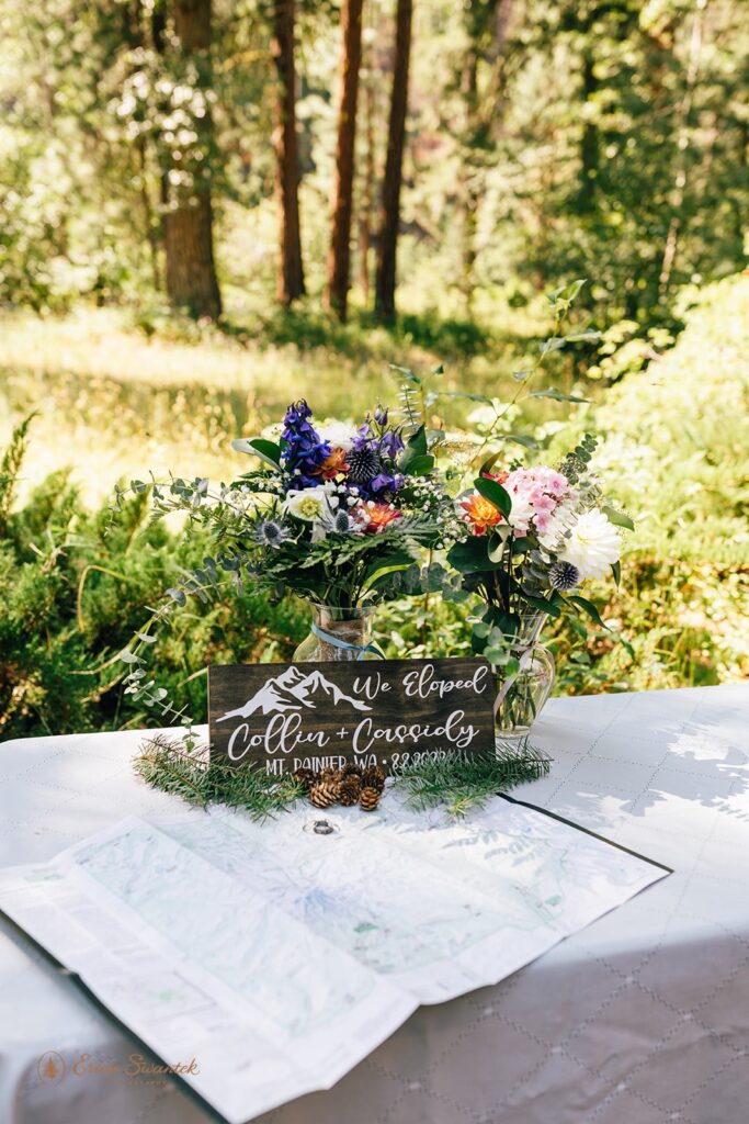 Two colorful floral bouquets in clear vases on a table with a map of Washington State, an elopement sign, pine cones, and wedding rings. 