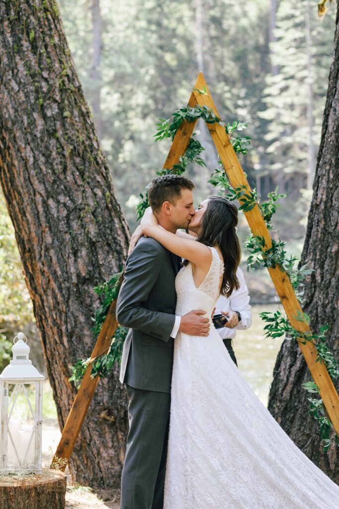 A couple kisses in front of an a-frame arch decorated with greenery during their intimate wedding at Whistlin' Jack's. 
