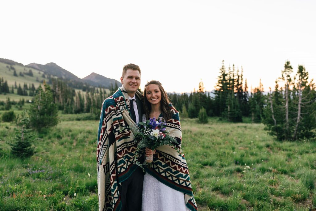 A hiking elopement couple poses for a wedding portrait wrapped in a printed blanket, holding a colorful bridal bouquet. 