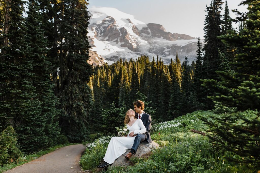 A woman in a long white wedding gown sits on a rock with her groom with Mt. Rainier in the background. 