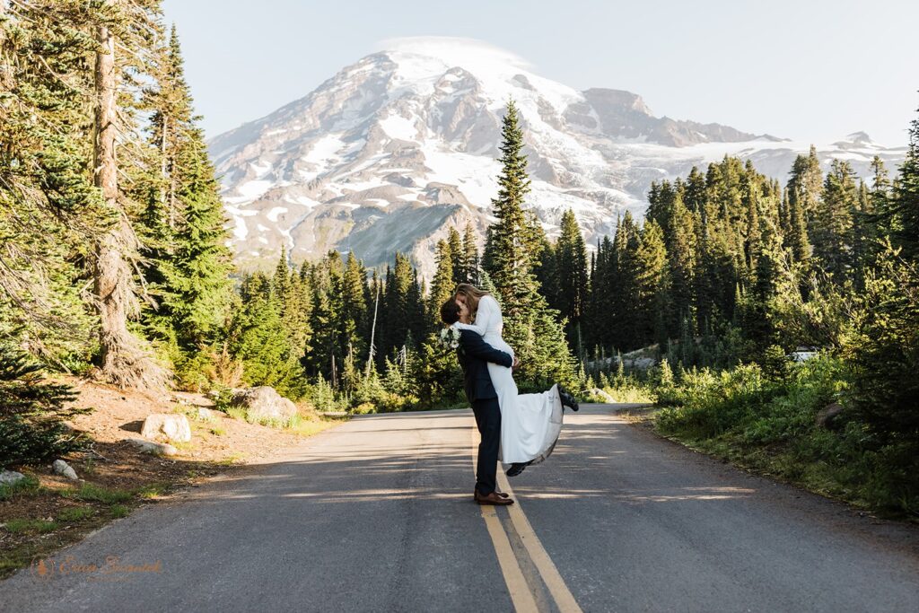 A groom lifts up his bride as they kiss on a road in Mt. Rainier National Park. 