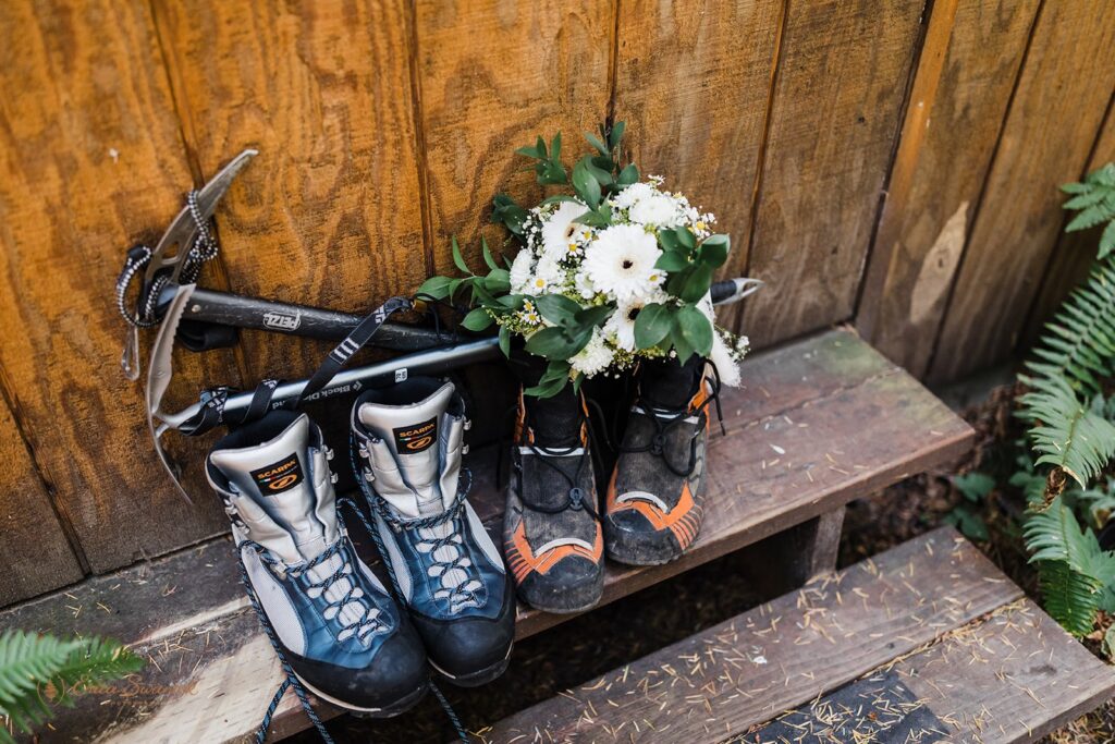 Two pairs of Scarpa Hiking boots sit on a wooden staircase while holding up two mountaineering axes and a white and green floral bridal bouquet. 