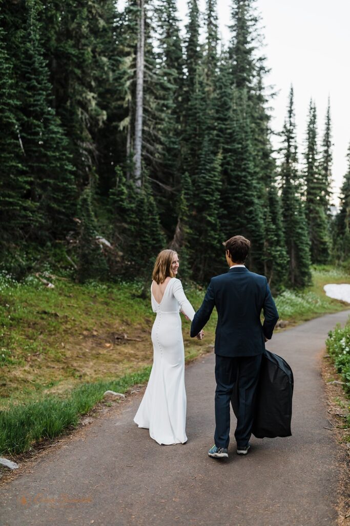 A Mt. Rainier elopement couple walks along a paved bath while the groom is holding a black garment bag. 