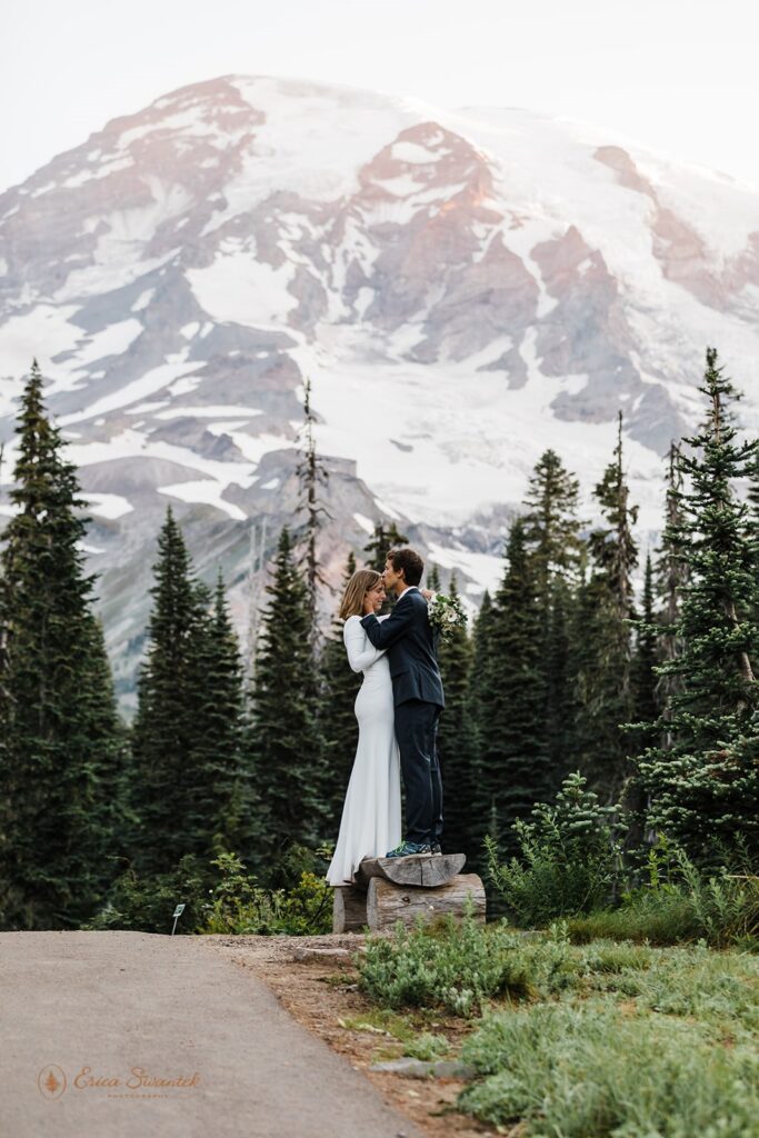 An adventure elopement couple holds one another while standing on a wooden bench with snowy Mt. Rainier in the background. 
