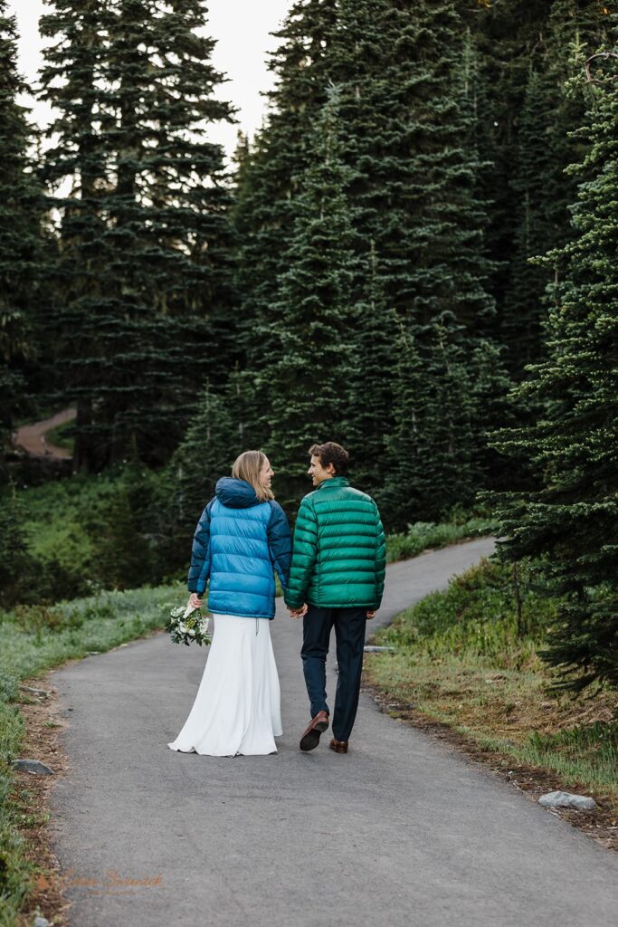 A hiking elopement couple holds hands along a Mt. Rainier National Park trail while wearing puffy Patagonia jackets. 