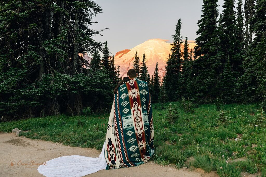 A Washington elopement couple cuddles underneath a printed blanket while admiring Mt. Rainier National Park at Sunrise. 