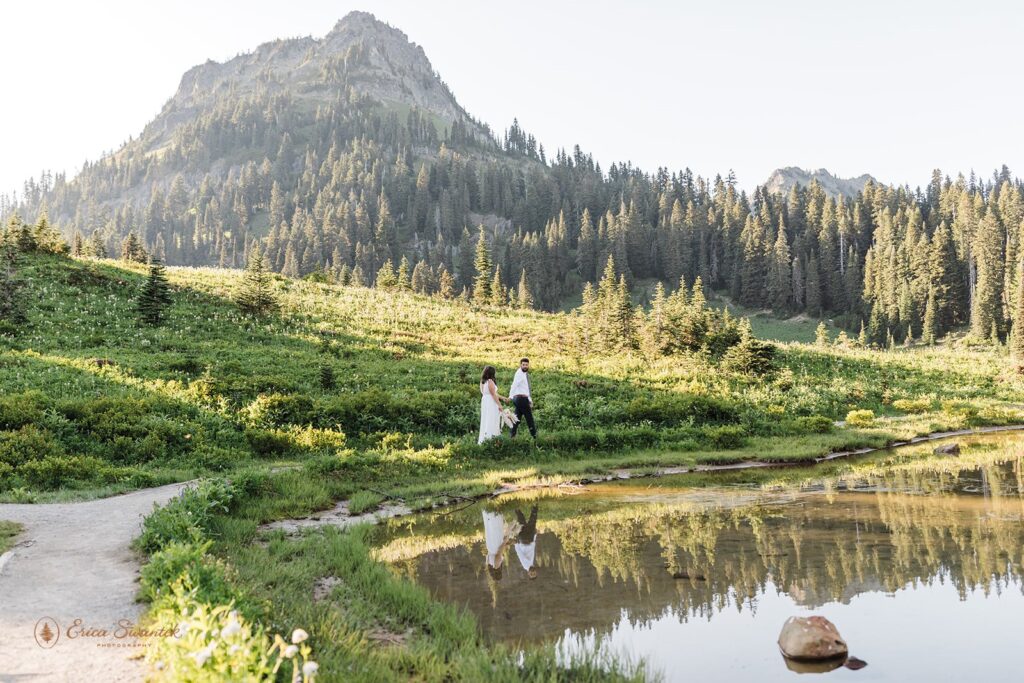 A couple in traditional wedding attire holds hands while walking around Tipsoo Lake. 