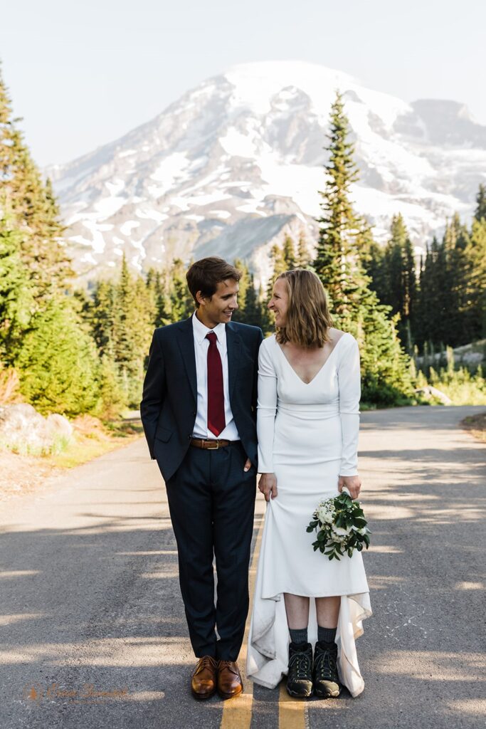 A groom in a suit and dark red tie stands with his bride along a road in Mt. Rainier National Park for wedding portraits. 
