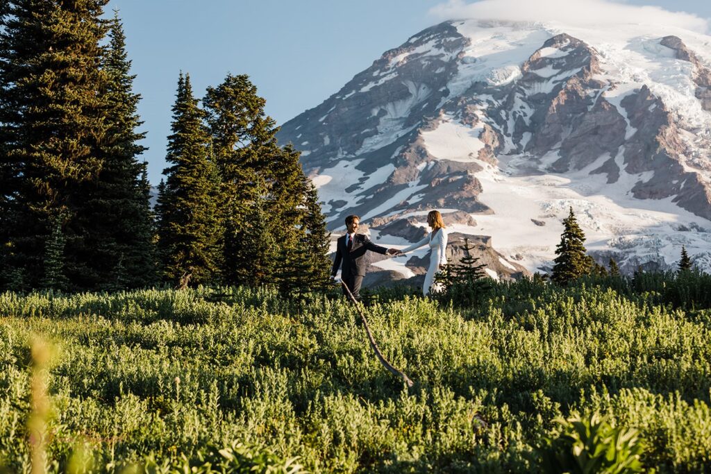 A hiking elopement couple strolls along a wildflower meadow trail in Mt. Rainier National Park while holding hands. 