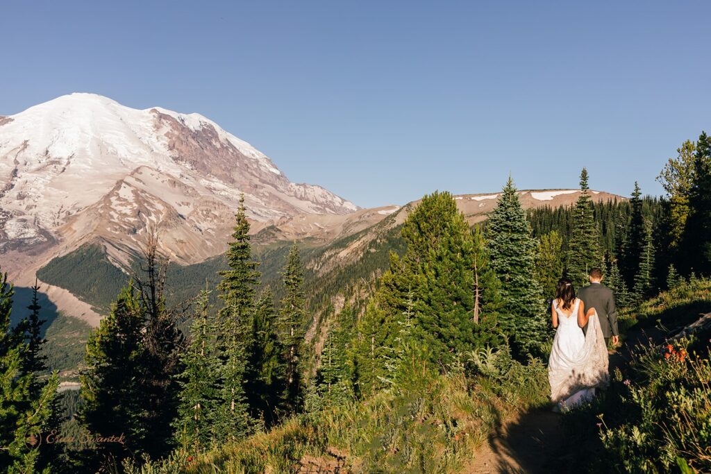A bride wearing a long, white wedding dress follows her groom in a suit during their Mt. Rainier hiking elopement at Skyline Loop. 