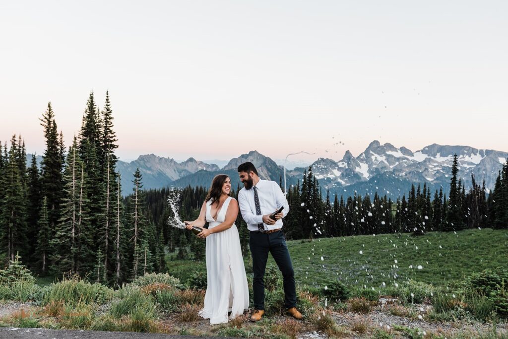 A couple wearing elopement attire celebrates their Summer Mt. Rainier National Park elopement by popping champagne at Sunset. 