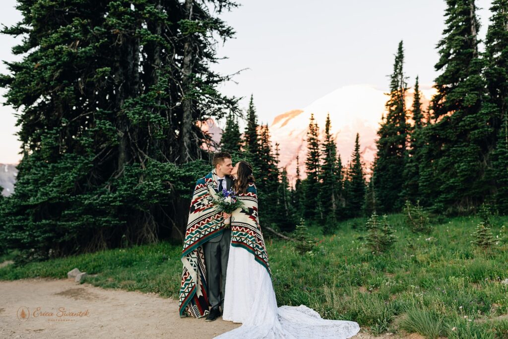 An hiking elopement couple wrapped in a printed blanket poses at Sunrise in Mt. Rainier National Park. 