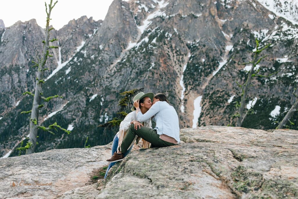A couple kisses as they sit on a large boulder in the North Cascades during their adventure couples session in Washington State. 